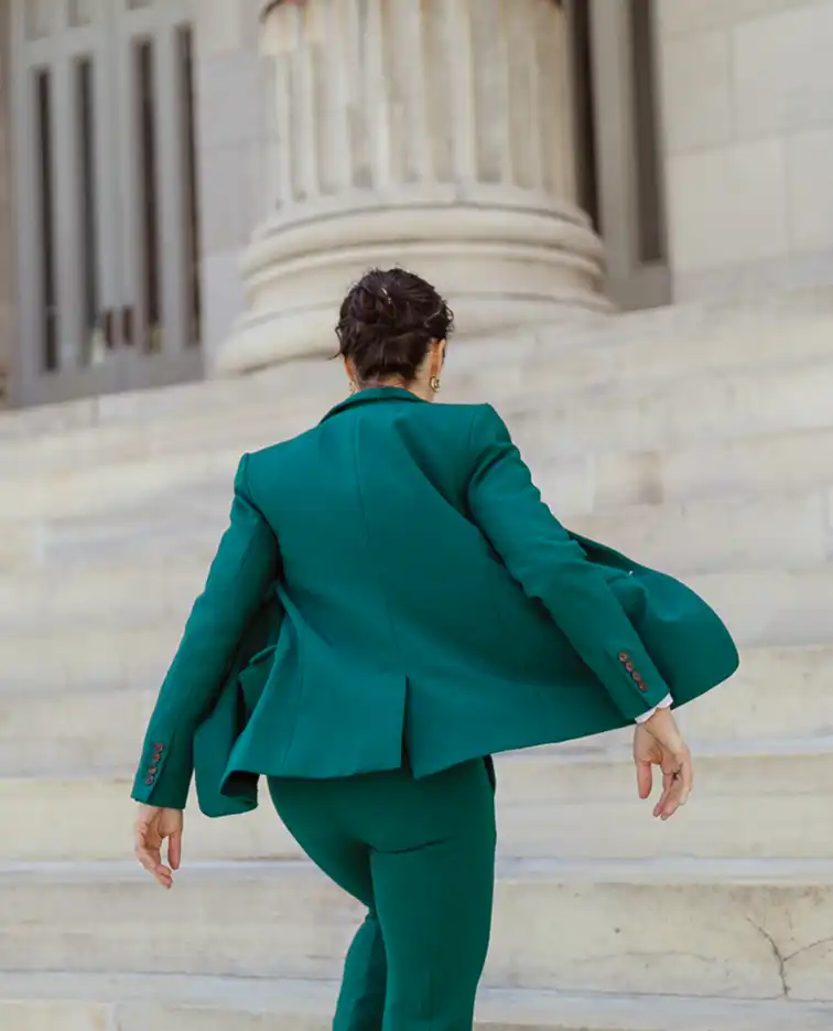Zoom-in view of Lina Stillman in a green suit ascends the stone steps of a large columned building, viewed from behind. A blue banner hangs above the entrance.
