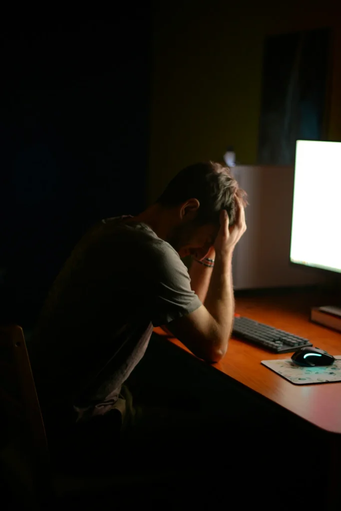 A person sits at a desk in front of a computer monitor with their head resting in their hands in a dimly lit room, conveying stress, fatigue, or frustration while working.