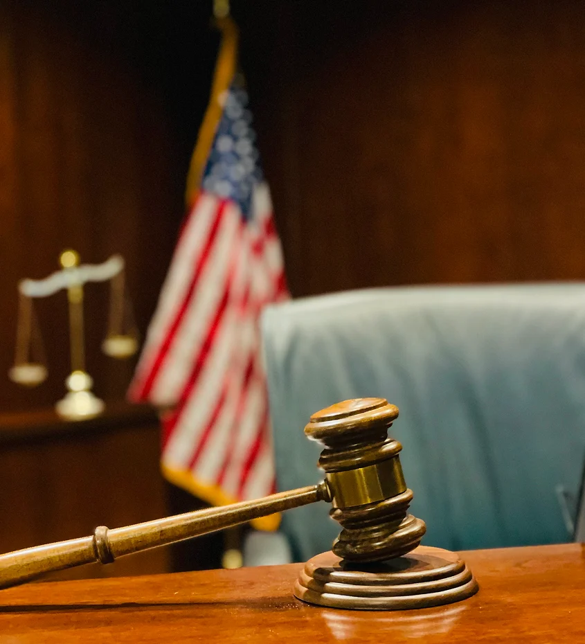 Wooden judge’s gavel on a courtroom bench with a blurred U.S. flag and scales of justice in the background.