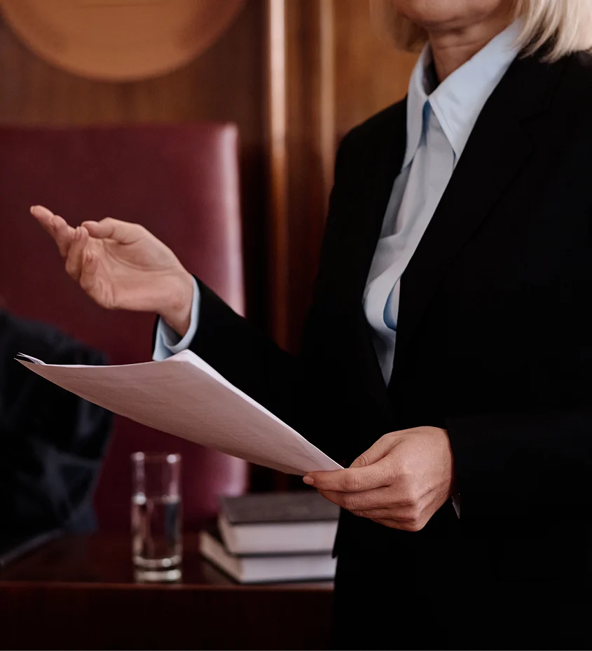 Attorney holding legal documents while speaking in a courtroom or legal hearing setting.