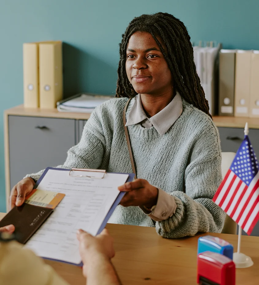 Black woman handing documents across a desk during an immigration or employment interview, with a small U.S. flag visible on the office desk.